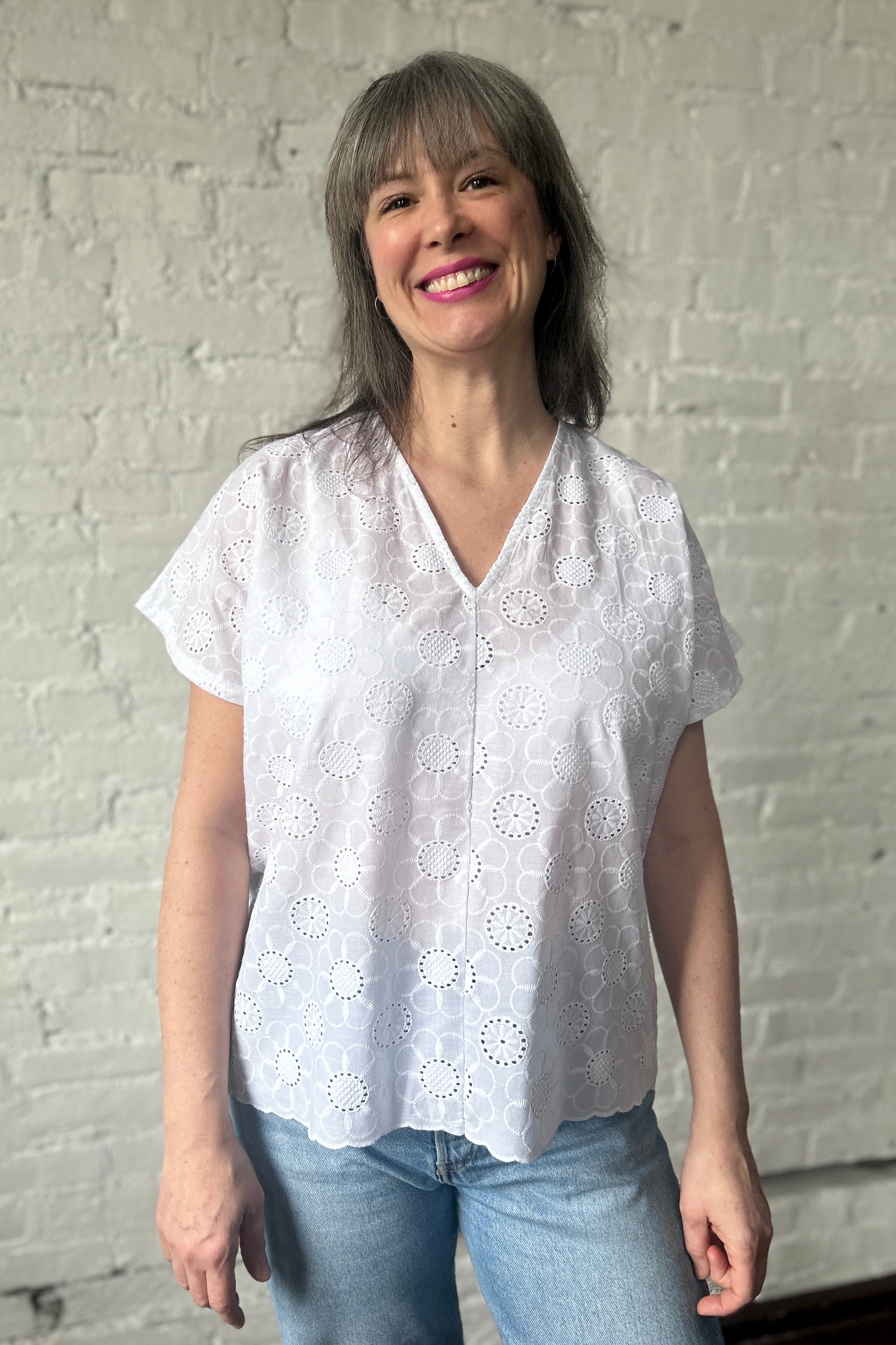 Smiling person models a boxy loose blouse in white cotton eyelet. Blouse has v-neck, short sleeves, and a scalloped hem. The eyelets are a large daisy pattern.