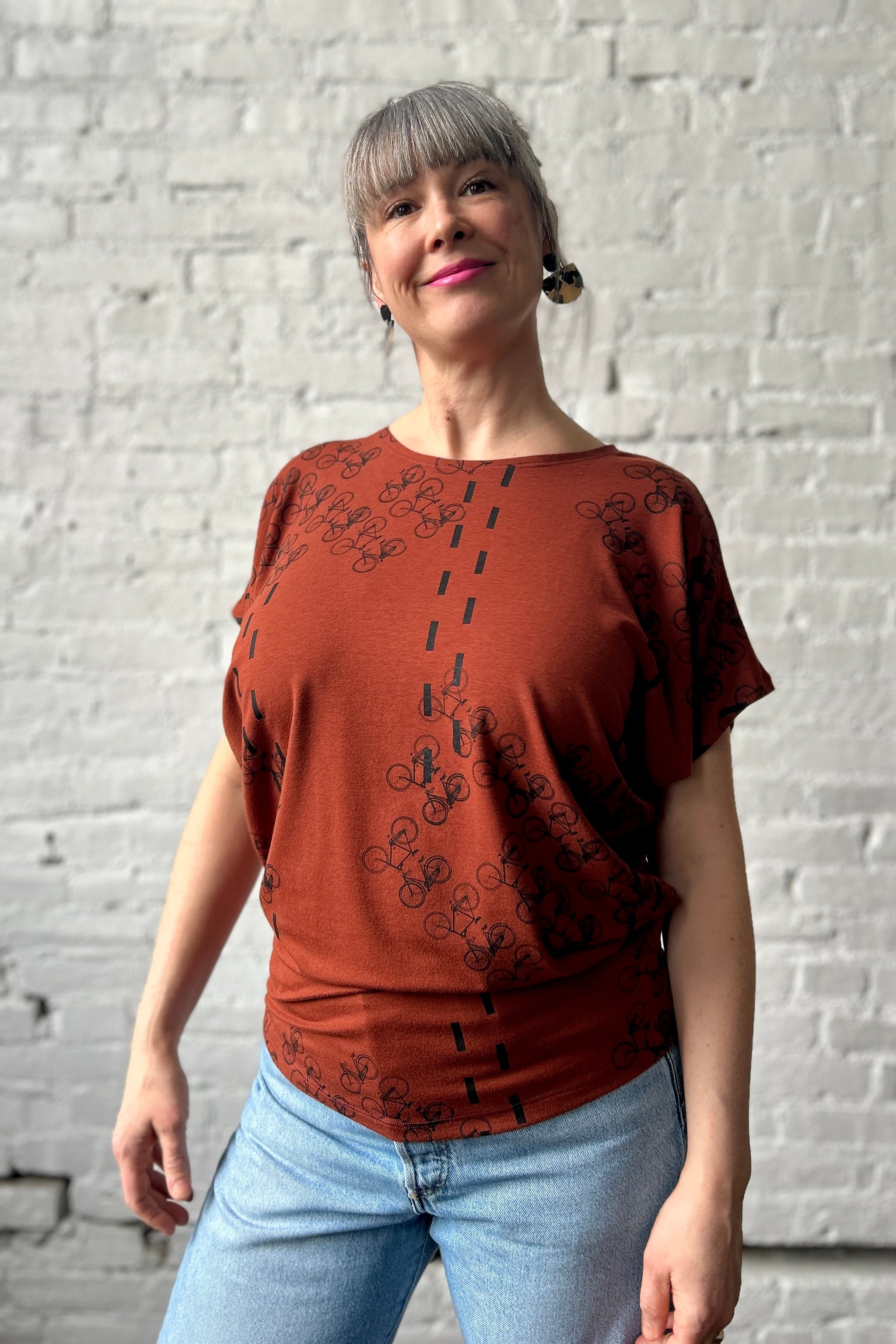 Woman wears a chestnut coloured drapey jersey-fabric short-sleevetop silkscreen-printed with black bicycles and paired with blue jeans.