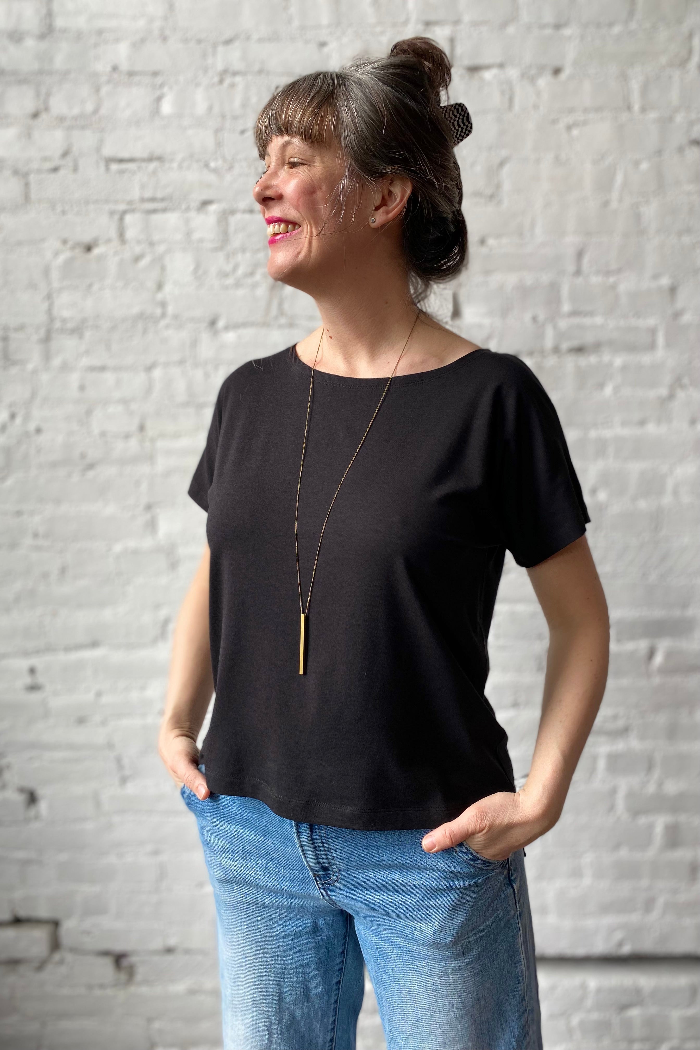 Woman wearing an unprinted black boatneck tee with gold necklace and blue jeans against a white brick wall background