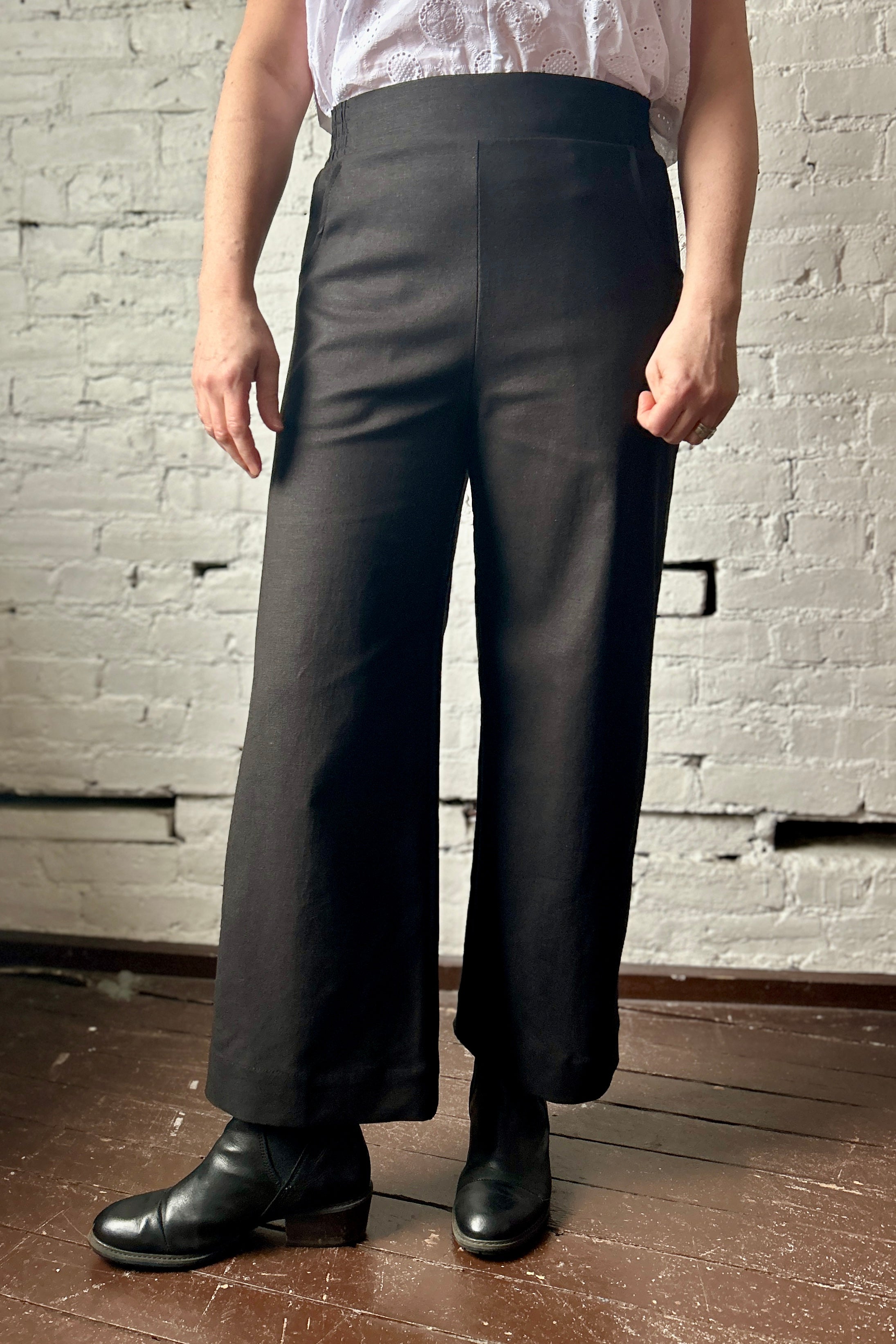 Person models black trousers with a flat front, cropped wide leg, side pockets, and elastic waistband. They wear black boots and a white blouse tucked into the waistband.