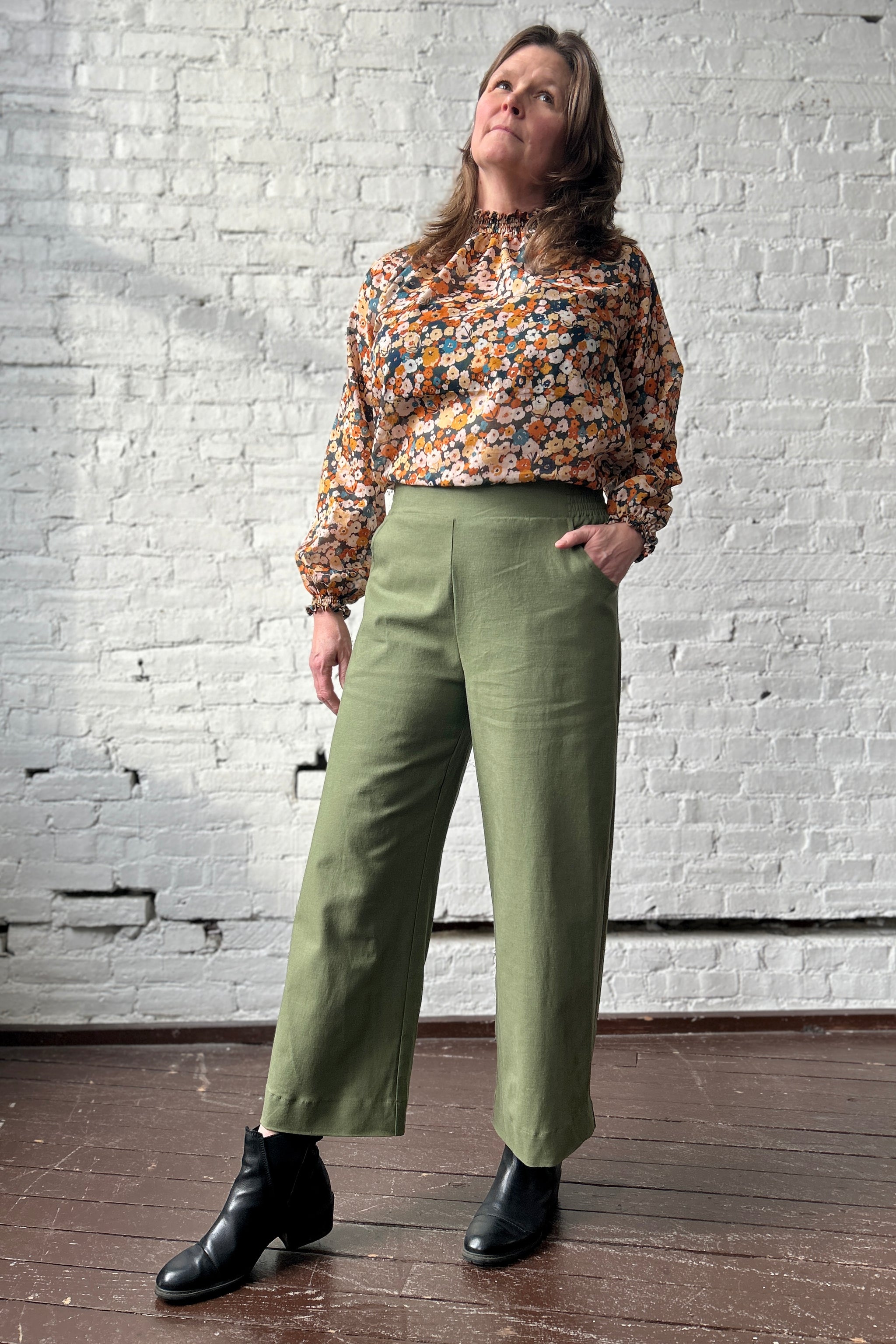 Person wearing olive-green, linen-blend trousers and floral blouse stands with hand on hip, with one leg forward. Trousers are high waist and wide leg with an elastic waistband and flat front.