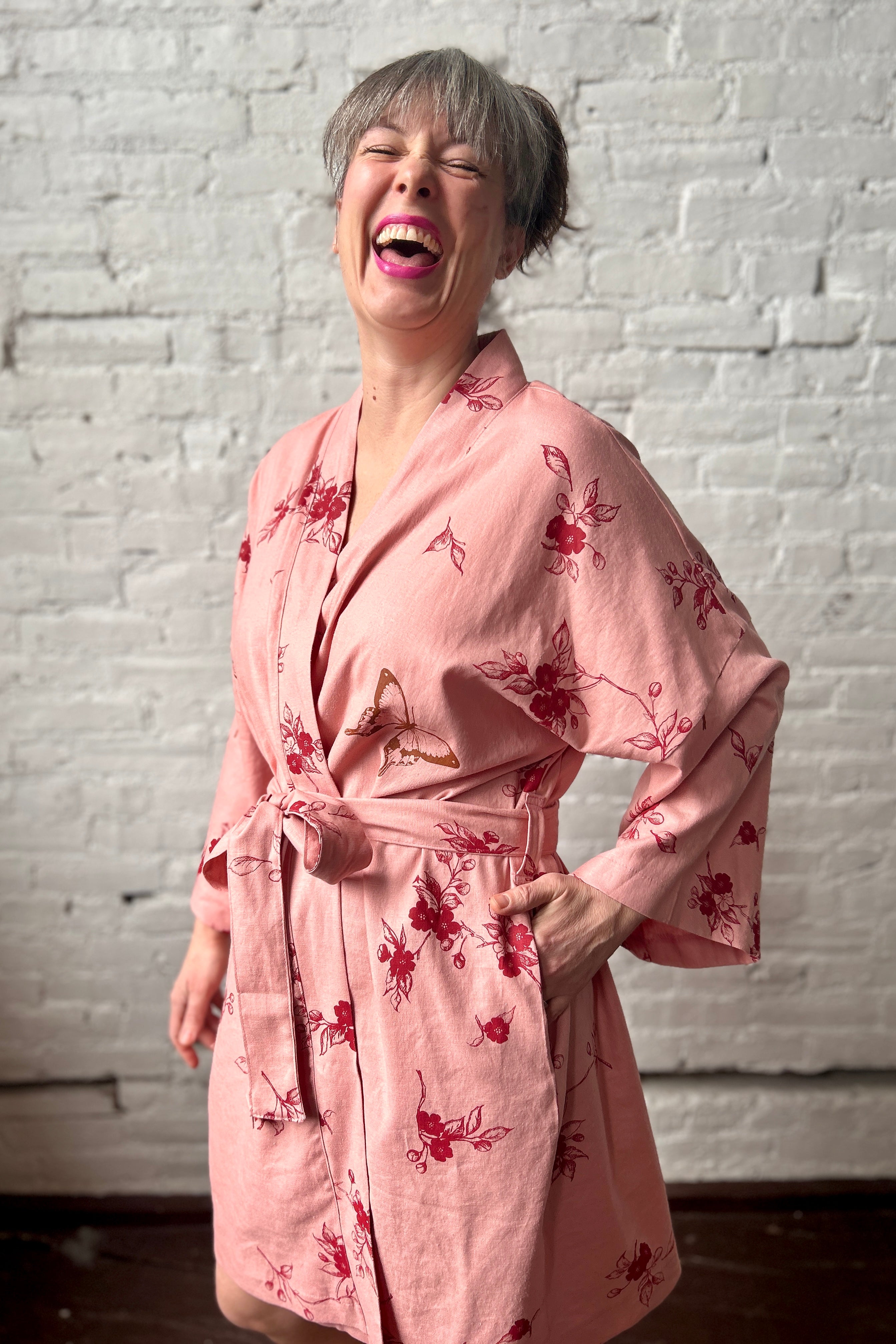 Laughing woman models a knee-length blush pink robe tied at the waist. Robe has loose sleeves, a long sash belt and large pockets. The fabric features a red apple blossom repeat print and various bugs printed in brown ink.