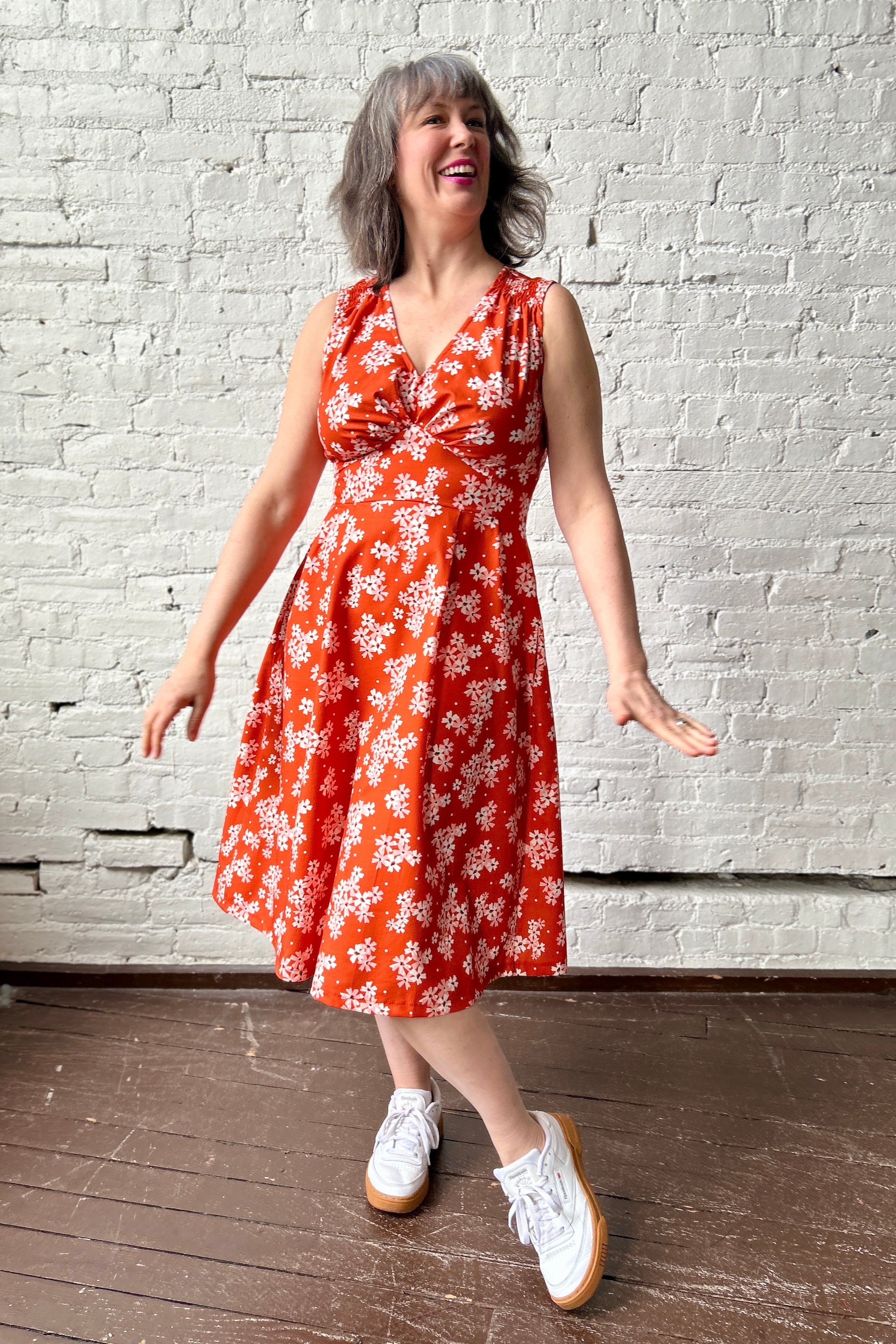 person models an orange sundress with print of white and pink small flowers. Dress has flared skirt, gathered shoulders and bust, fitted waist band.