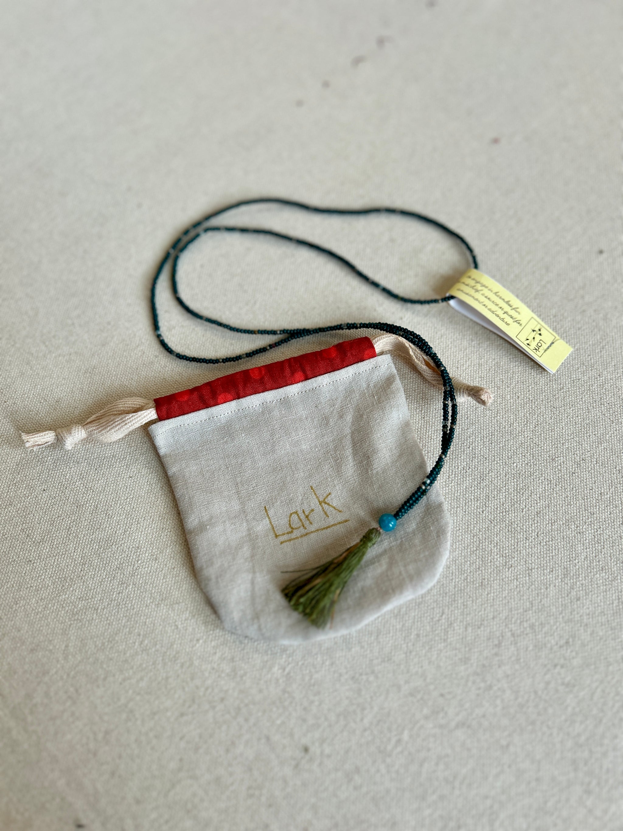 A beaded necklace with tassel lies coiled over a fabric bag with the word 'Lark' printed on it.
