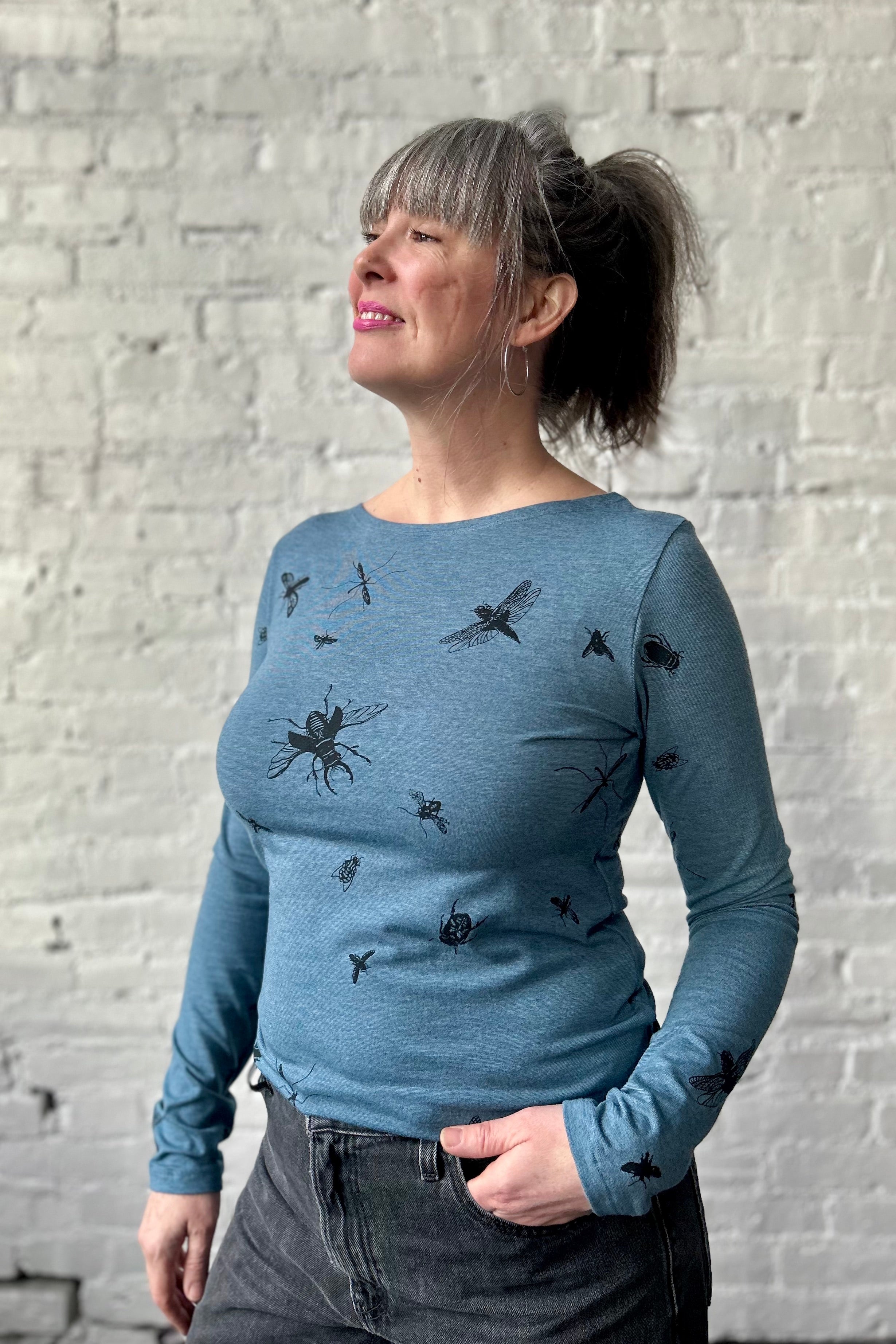 Smiling woman stands with a hand in pocket, modeling a blue microstriped long sleeve boatneck shirt. The shirt has black silkscreened bugs printed in on it in a large repeat pattern.