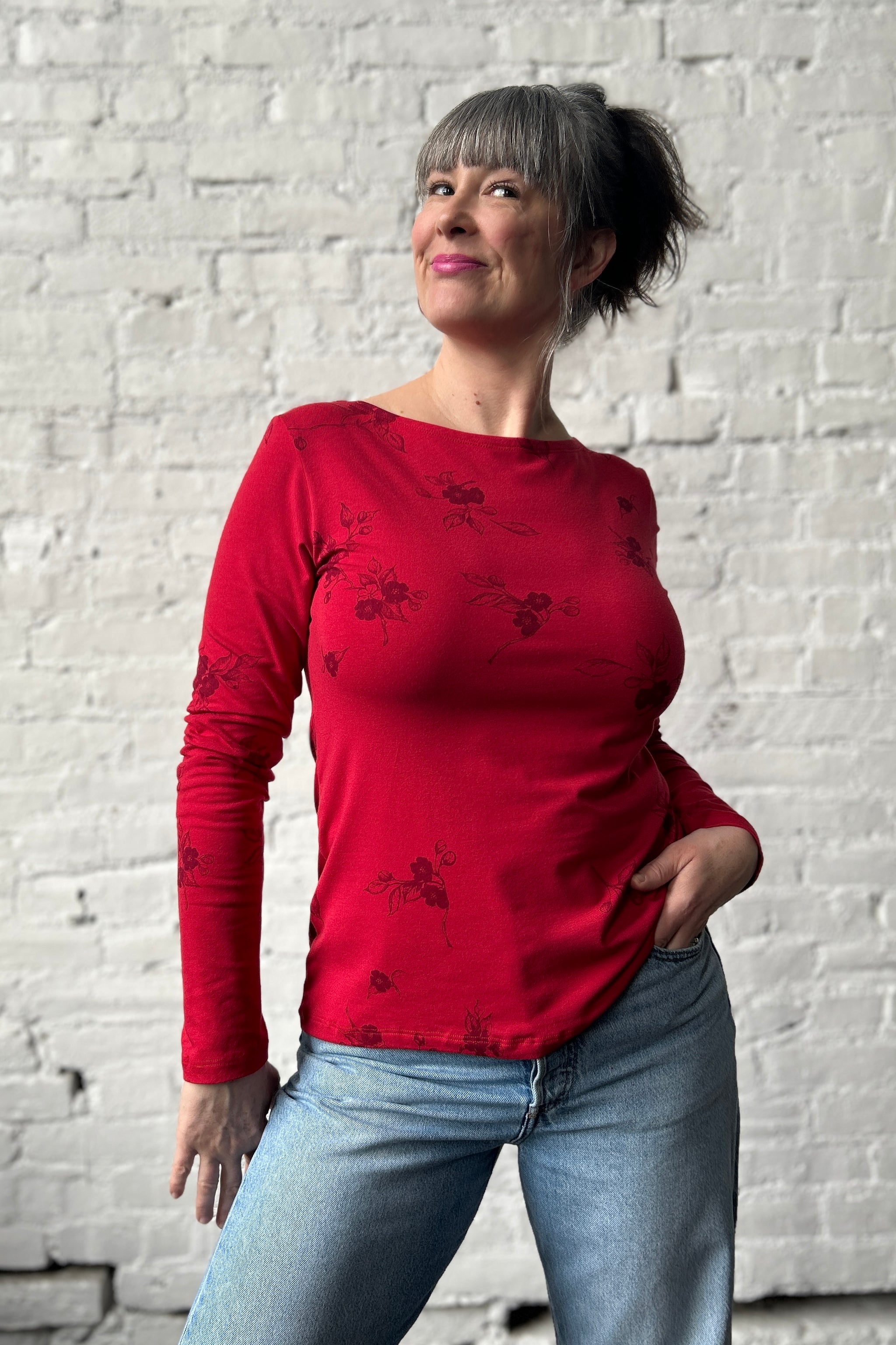 Woman poses with hand in jeans pocket, wearing a long sleeve red tee with boatneck. Shirt features apple blossom repeat print in dark red ink.