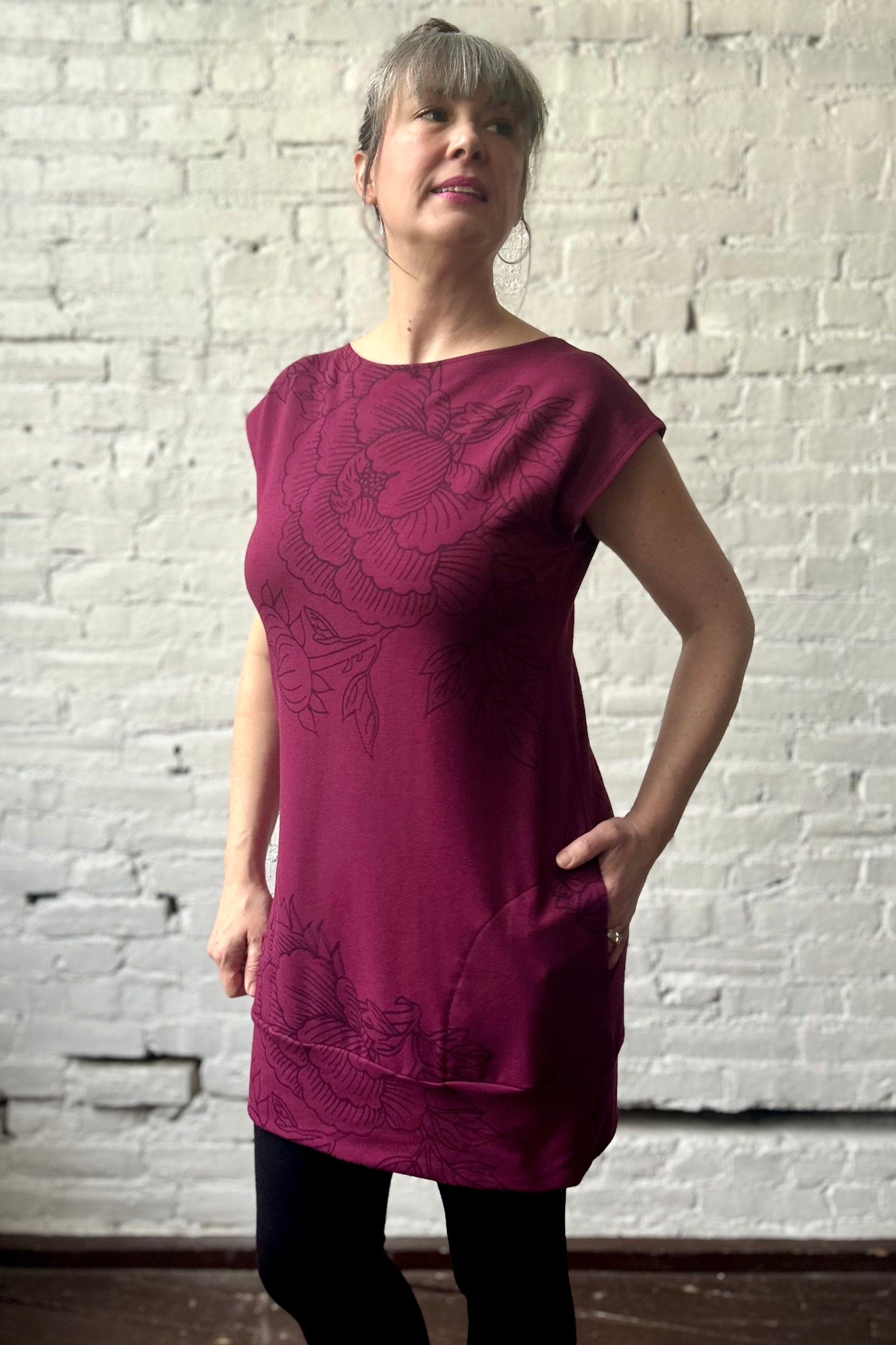 Woman wears a wine-coloured cap-sleeved tunic with giant peony outlines printed on it a large repeat pattern. Worn with black leggings