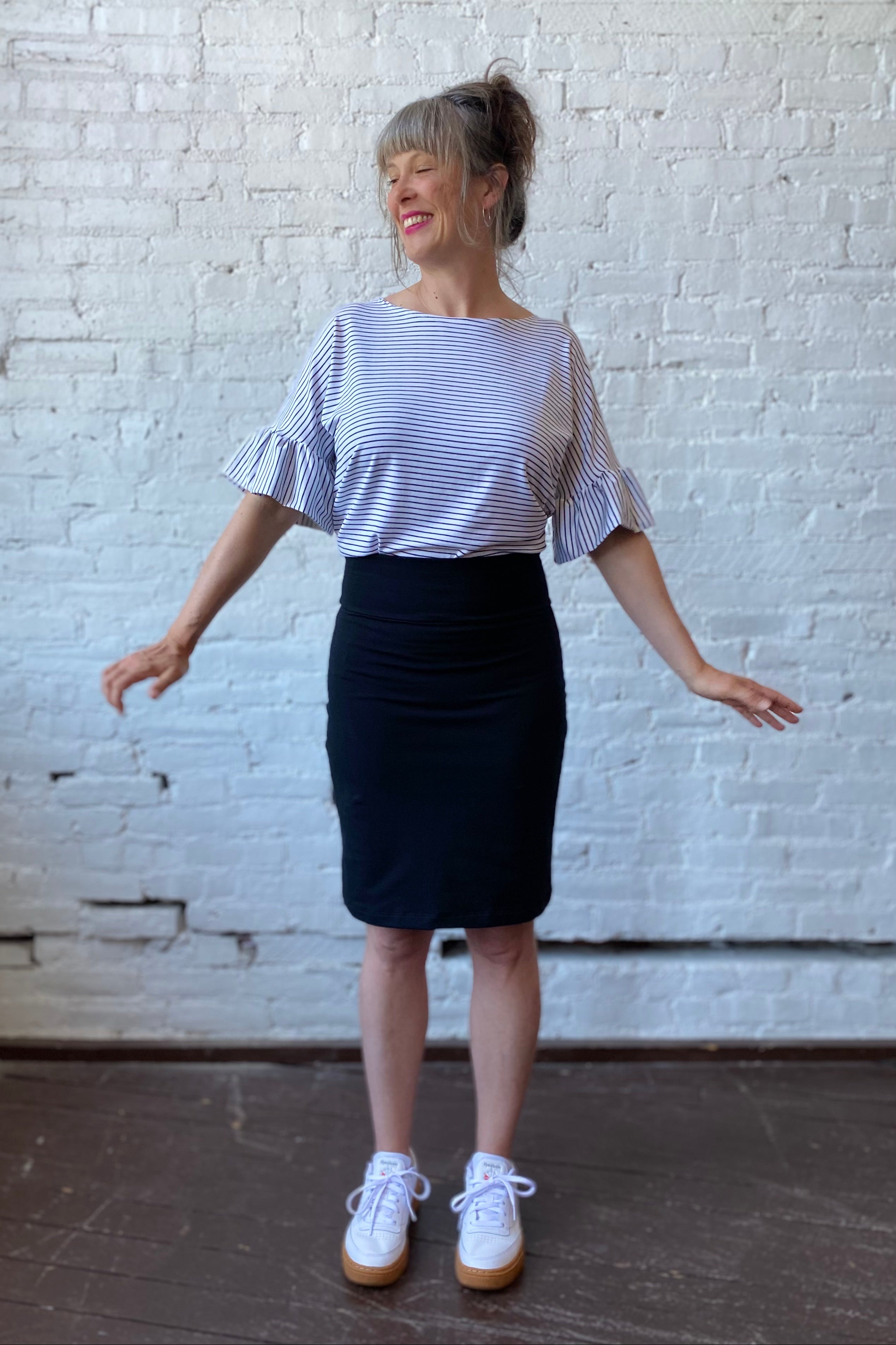 Standing woman wears a knee-length black pencil skirt with striped blouse and white sneakers 