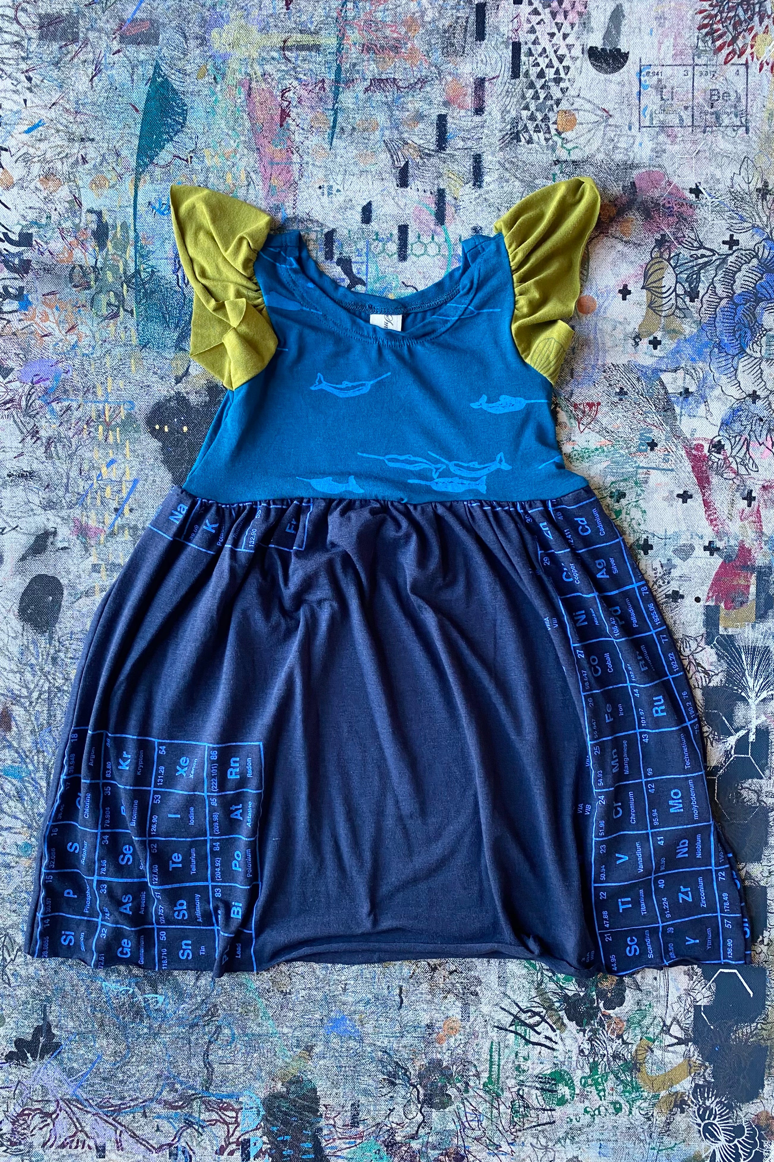 Kid's dress laid flat, with ruffled cap sleeves and gathered skirt. Torso, skirt and sleeves are in different colours of jersey fabric with silkscreen patterns in various designs. 