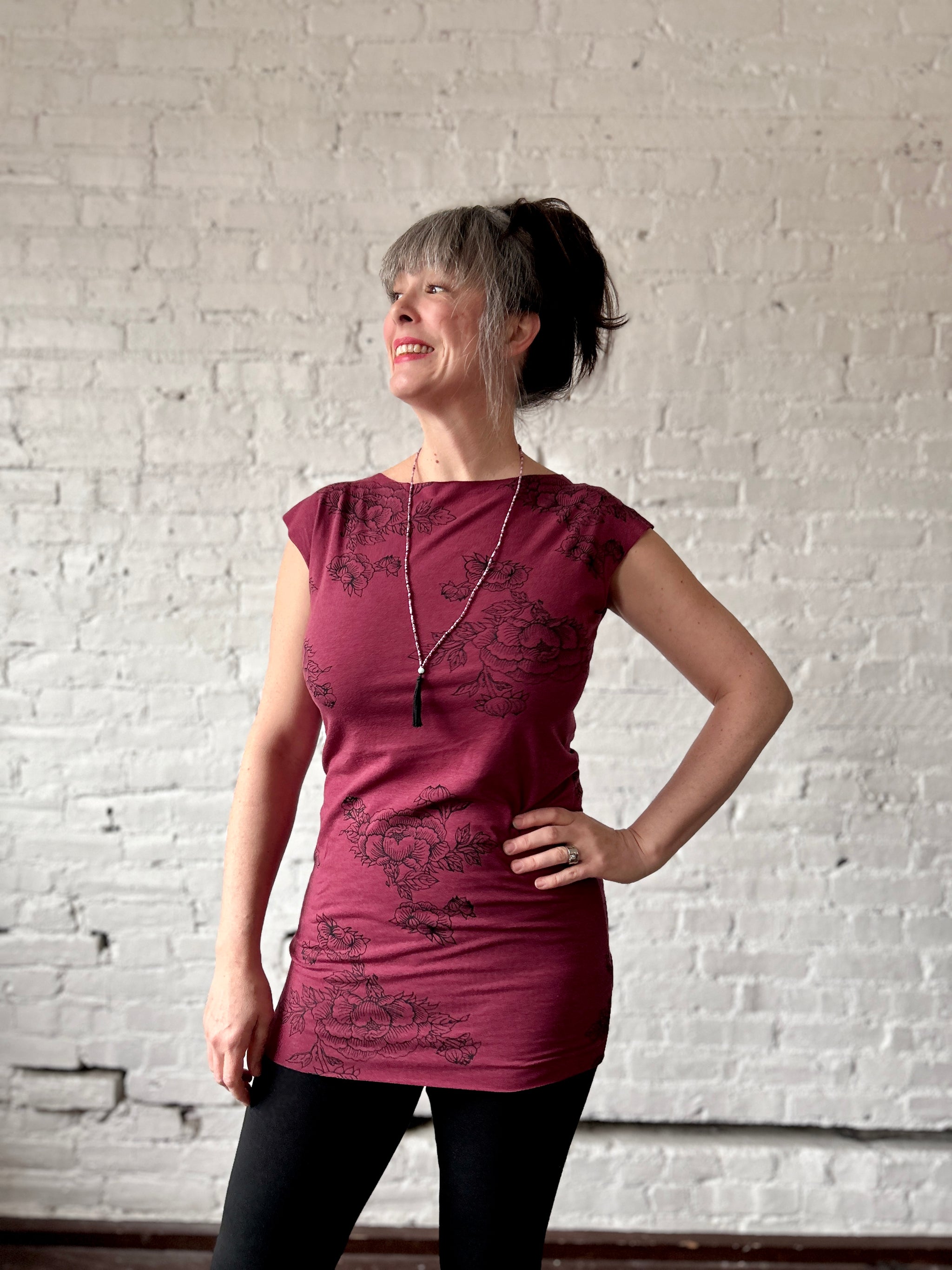 Smiling woman stands with hand on hip in a long burgundy maroon tee shirt printed with black peony outlines. 