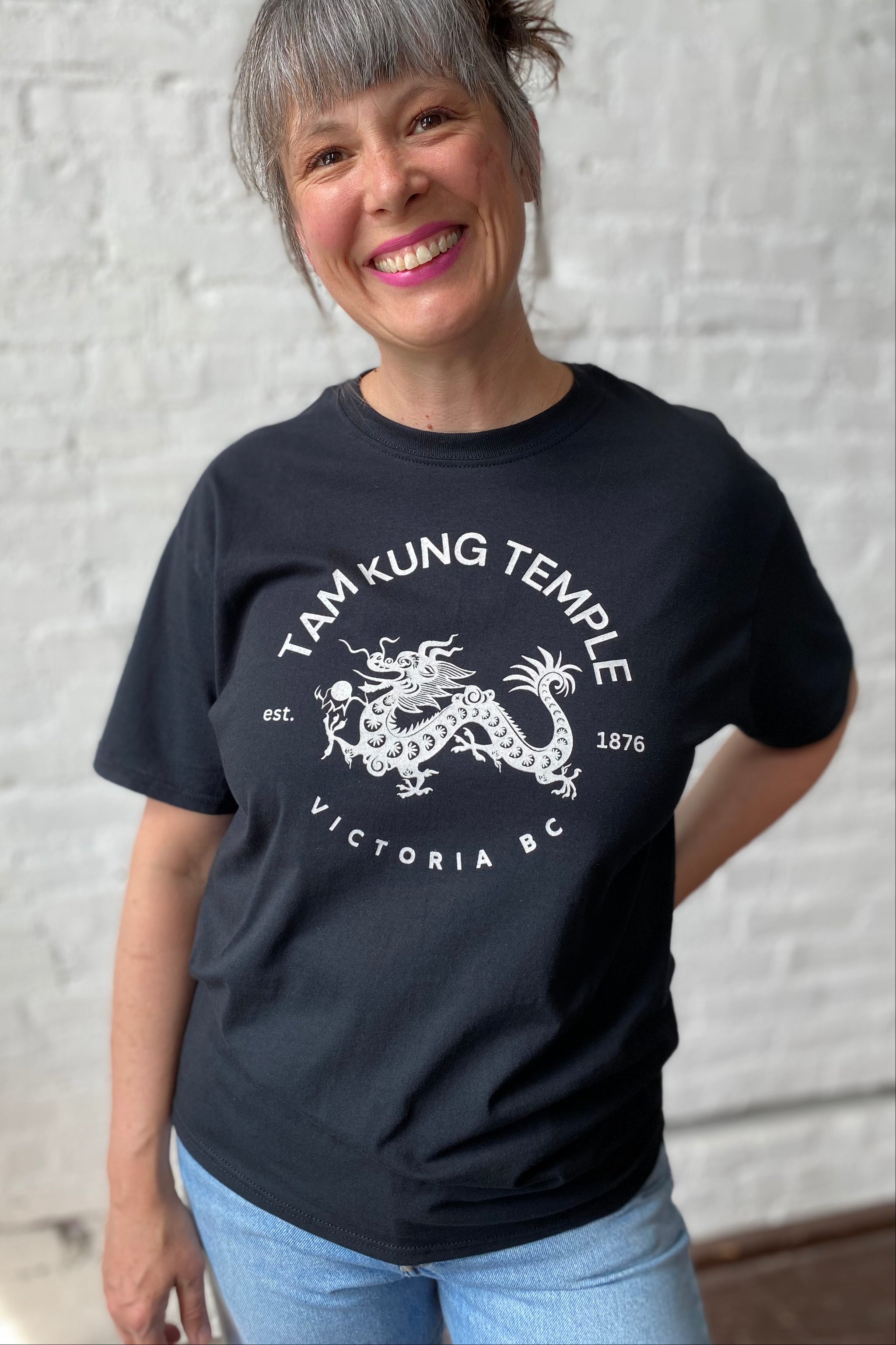 Smiling woman models black unisex tee with a white chinese dragon printed on chest. Dragon is encircled by words 'Tam Kung Temple est. 1876 Victoria BC'