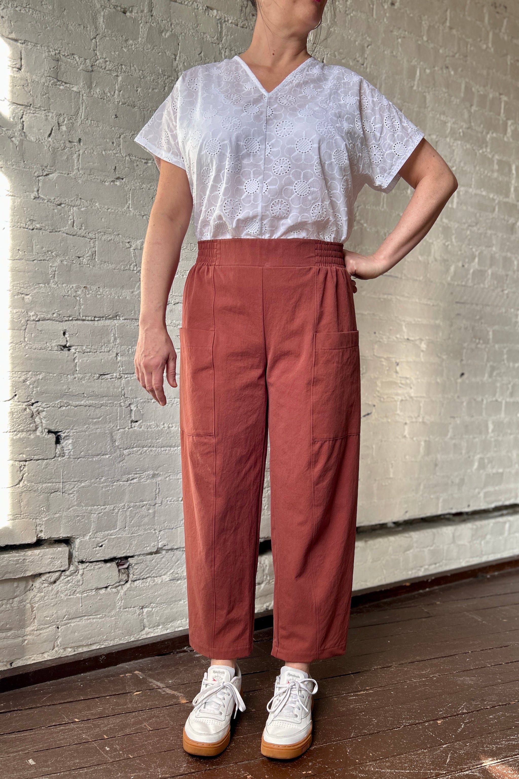 Person wears red-clay coloured cotton trousers and white eyelet blouse, standing with hand on hip. Pants have front seam, barrel leg, elastic waist with flat front, and deep utility pockets on front-side of legs.